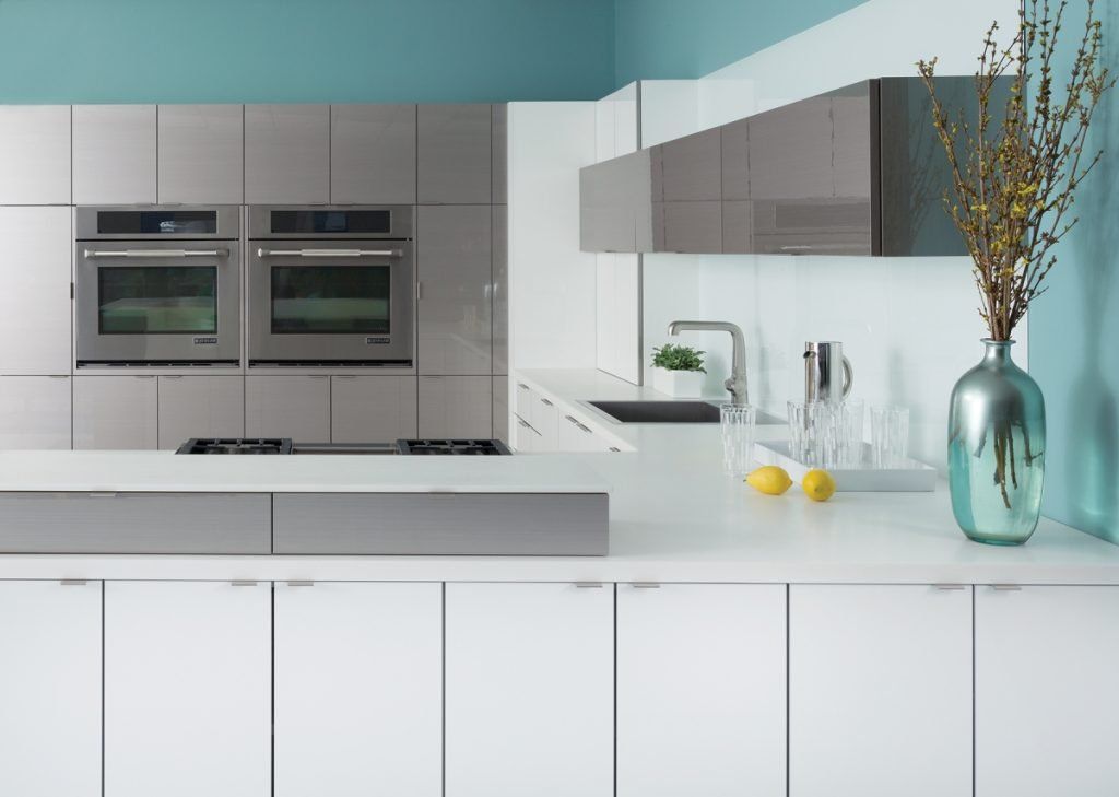 Modern white kitchen with built-in ovens, gray cabinets, and a white countertop with a vase and fruit.