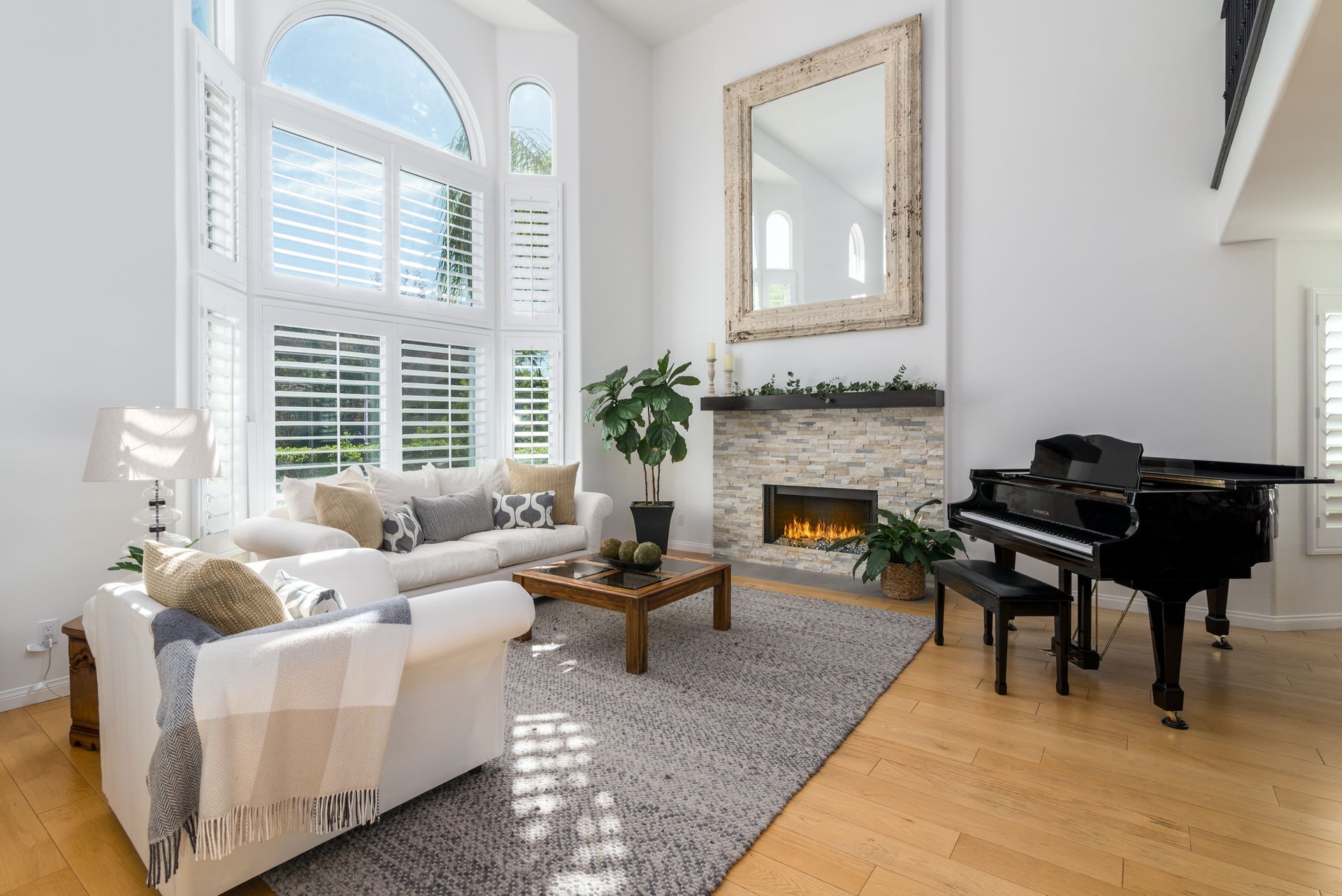 Bright living room with tall windows, fireplace, white sofa, and a black grand piano on hardwood floors.
