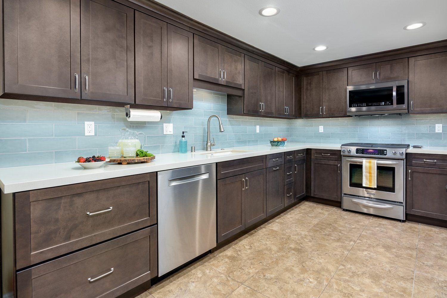 Modern kitchen with dark cabinets, light blue backsplash, stainless steel appliances, and beige tile floor