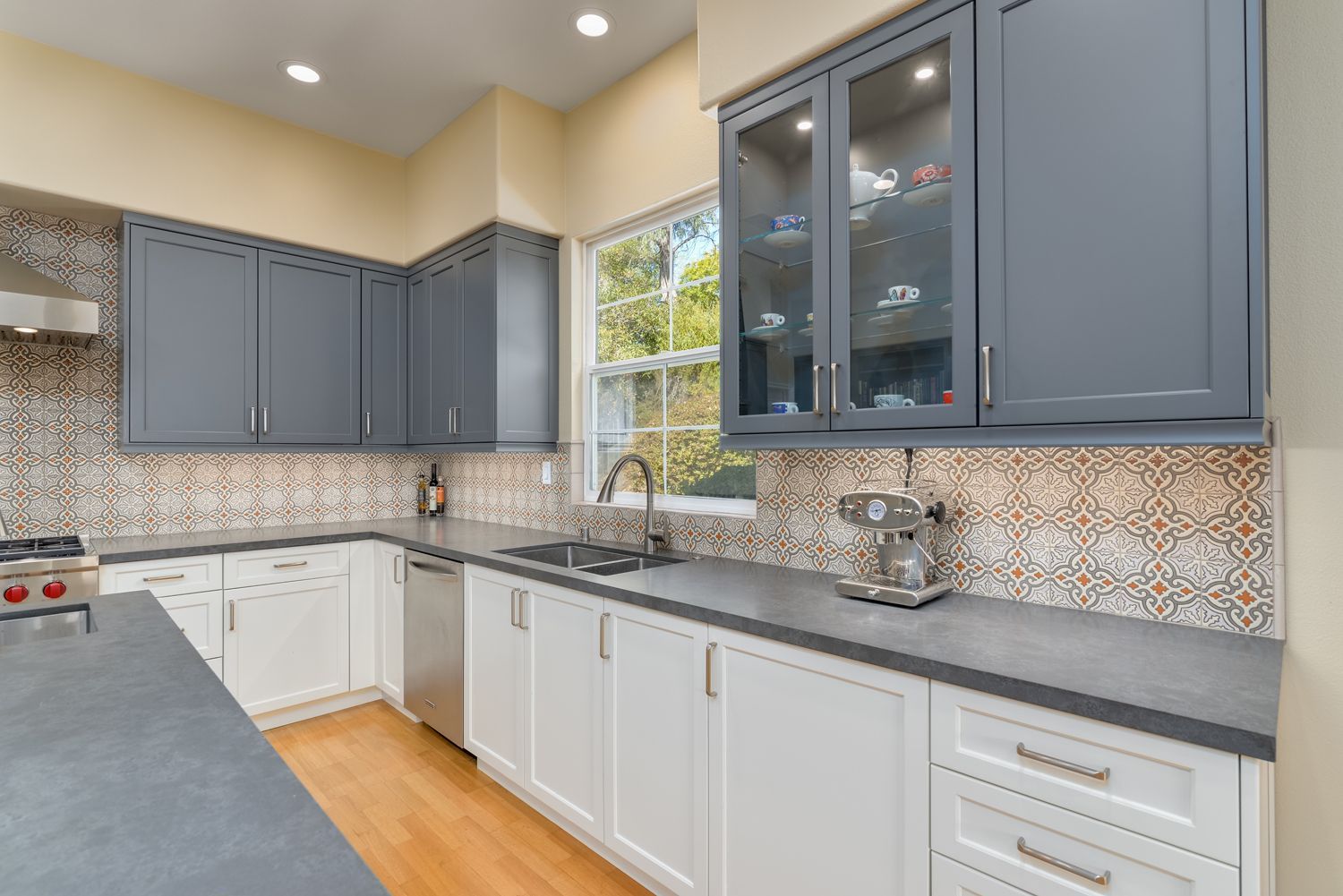Modern kitchen with blue-gray and white cabinets, gray countertops, patterned backsplash, and a window corner.