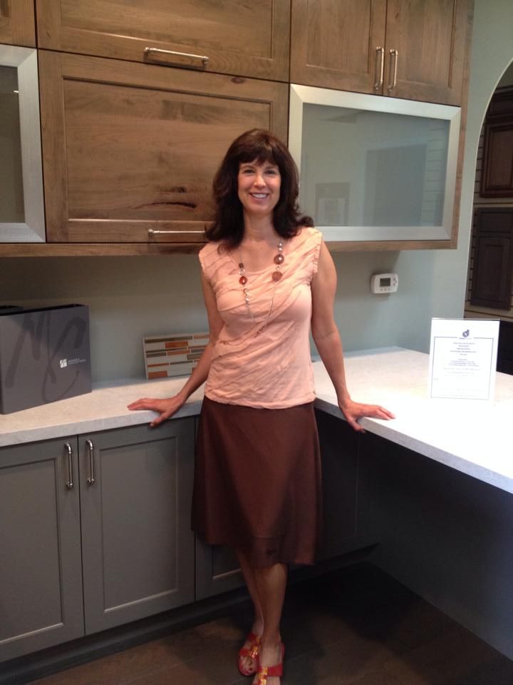 Woman standing in a kitchen, leaning on a white counter, wearing a pink top and brown skirt.