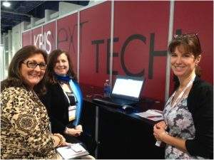 Three people smiling at a tech booth with a laptop and red wall backdrop