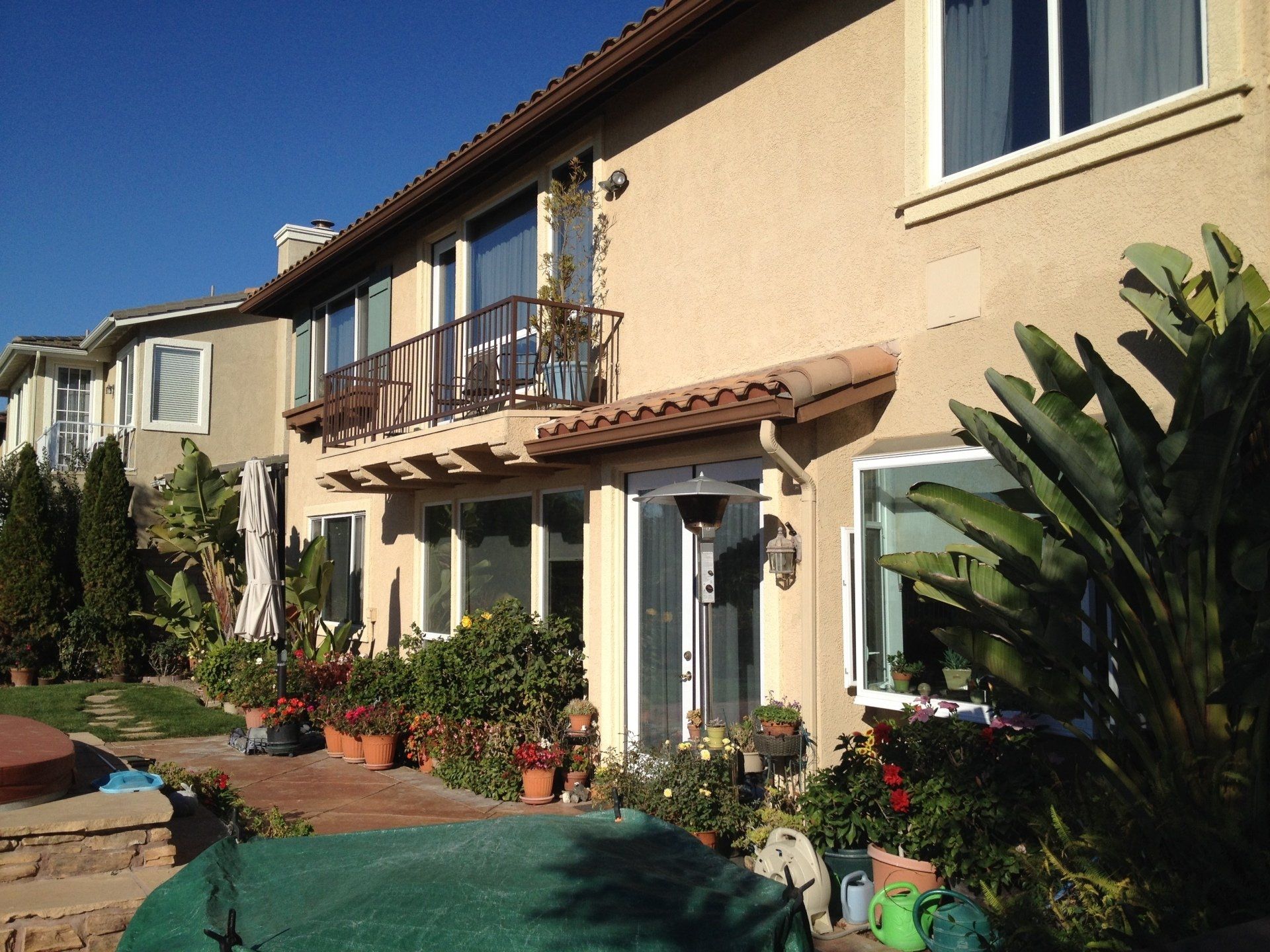 Sunlit stucco house with balcony, potted plants, and a landscaped front yard under a clear blue sky