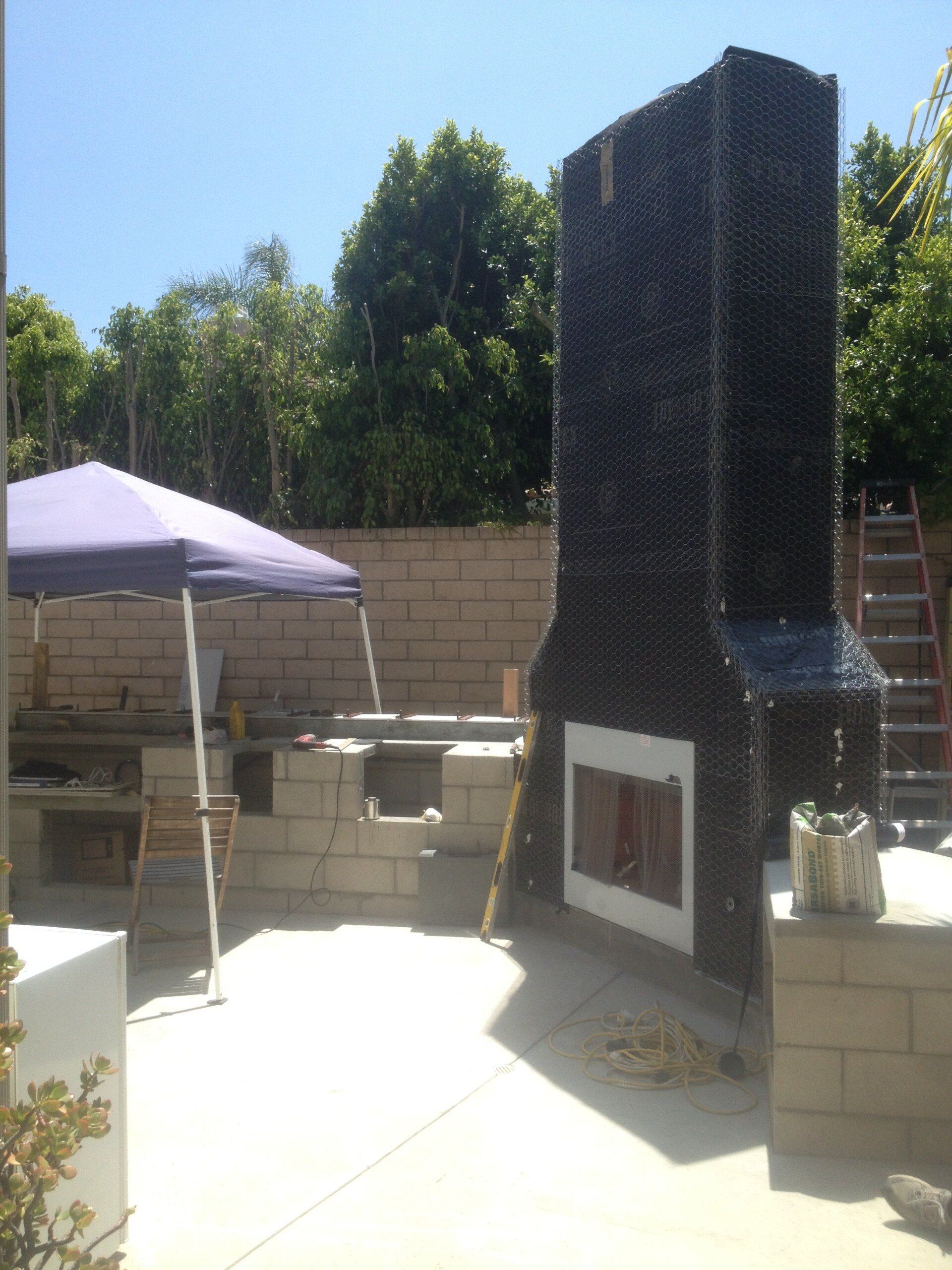 Backyard patio with a tall black outdoor fireplace, white tent canopy, and cinderblock seating in sunlight