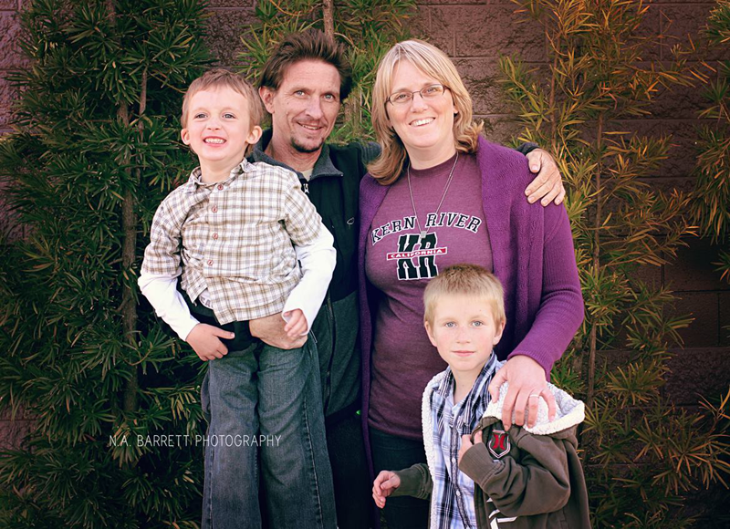 Family of four posing outdoors by a brick wall, smiling and standing close together.