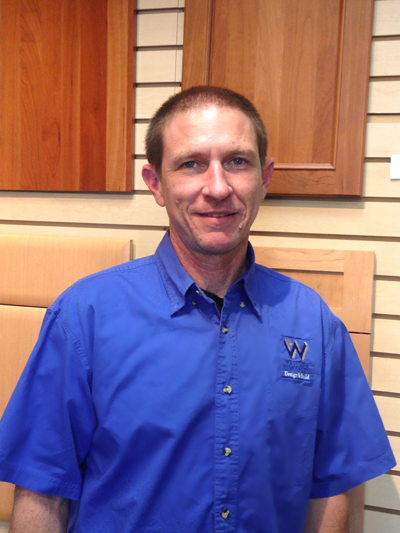 Man in blue button-up shirt standing in a wood-paneled room, smiling at the camera.