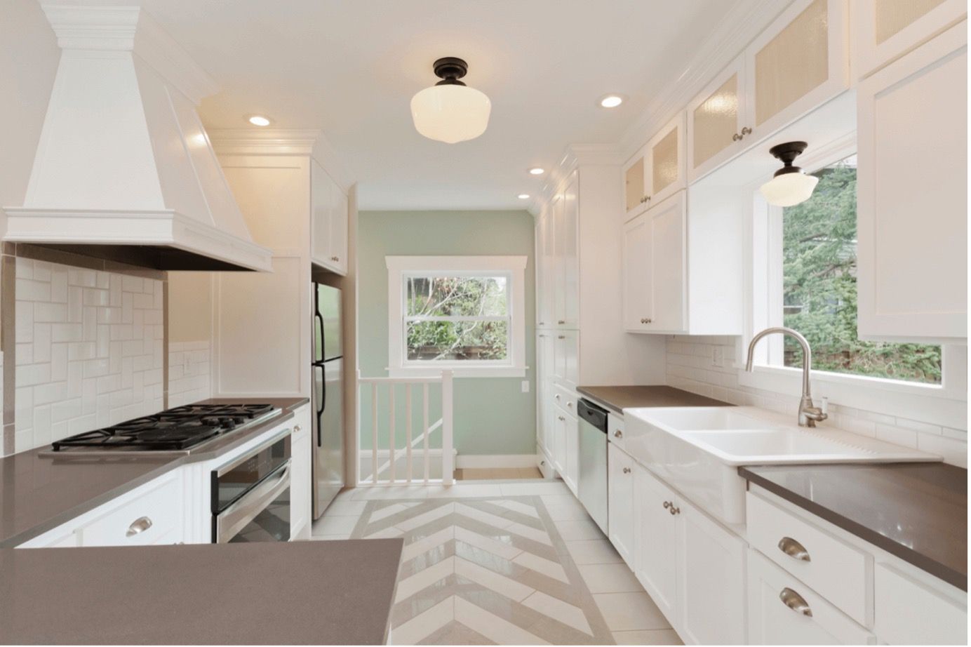 Bright galley kitchen with white cabinets, dark countertops, chevron floor, and a window over the sink