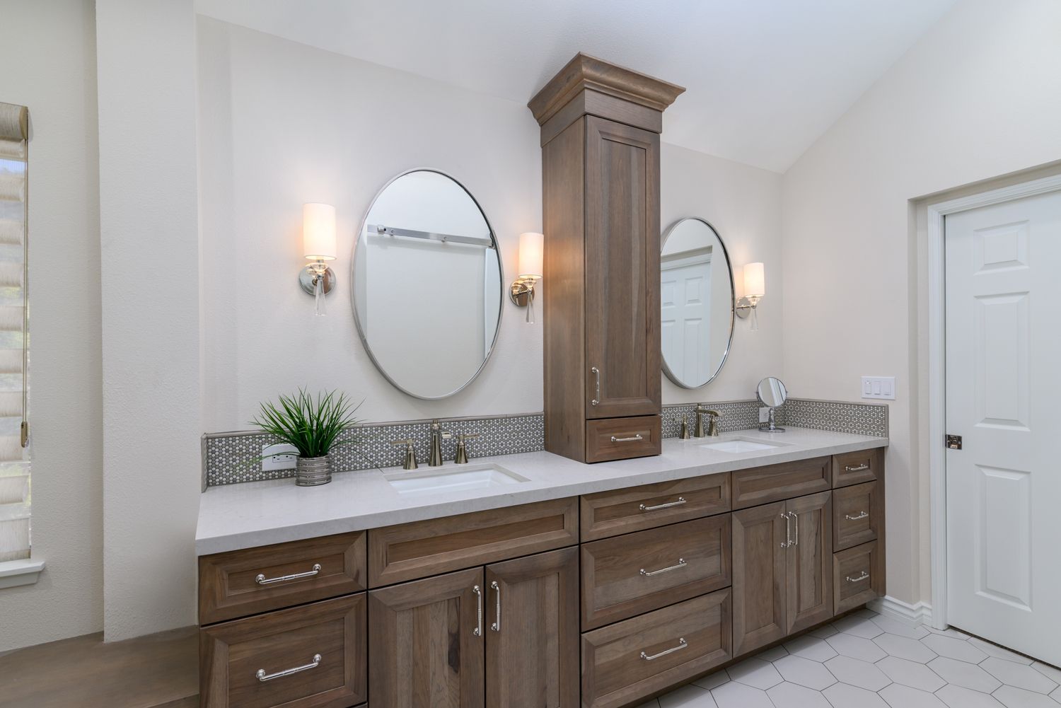 Elegant bathroom vanity with double oval mirrors, wall sconces, and wood cabinets beneath a marble countertop