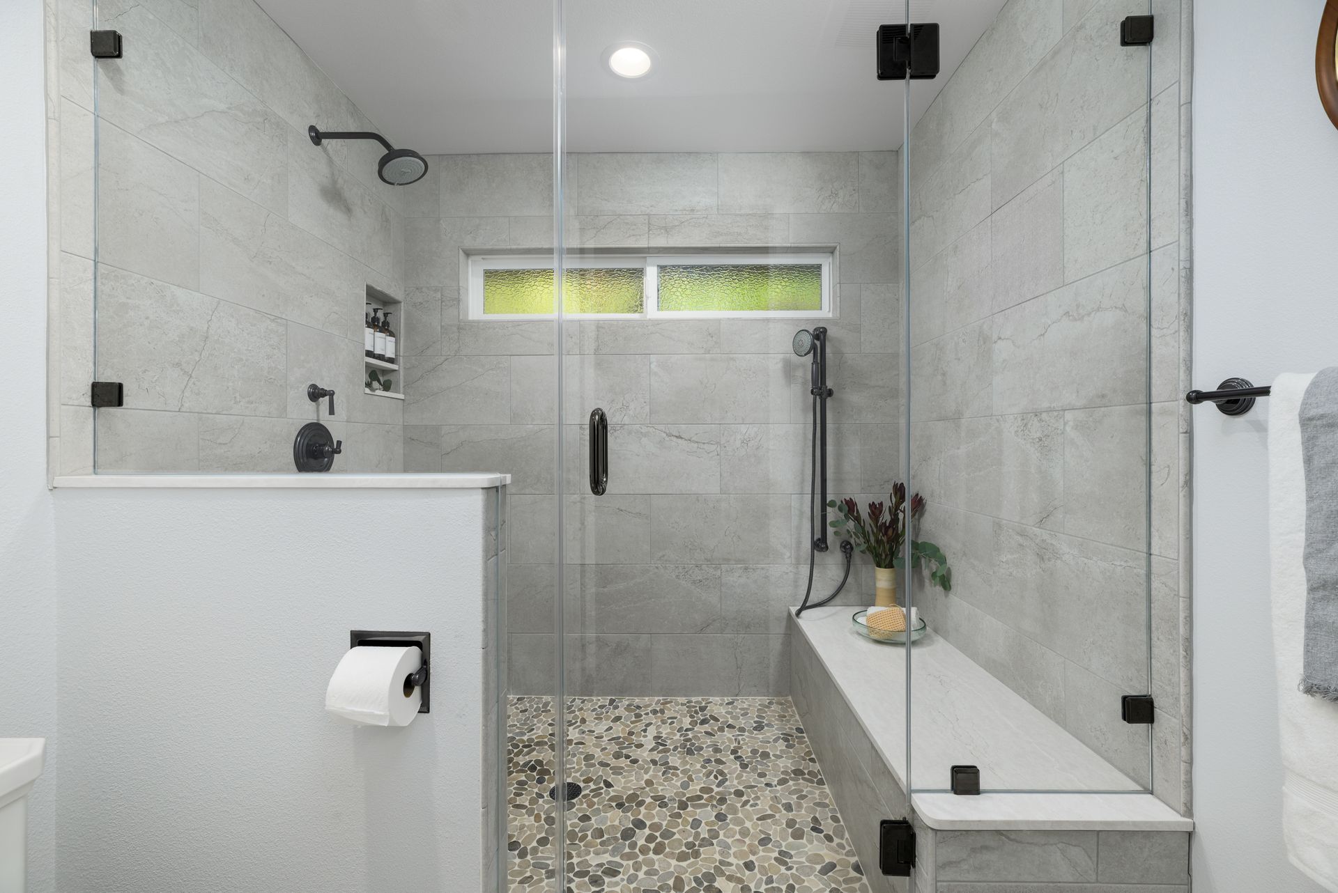 Modern white tiled bathroom with a walk-in shower, glass partition, and pebble tile floor.
