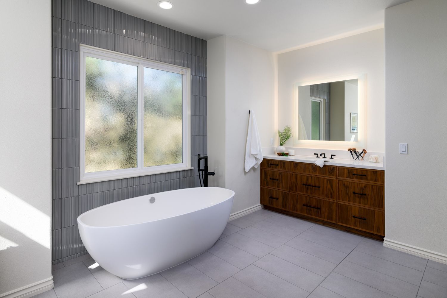 image shows a bathroom with a tub under a window, a walnut vanity and lighted mirror, Thousand Oaks,CA primary bathroom remodel 