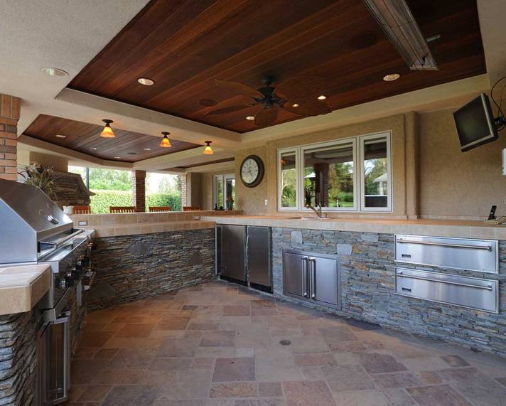 Outdoor kitchen with stone counters, stainless appliances, and a wood-paneled ceiling under a covered patio