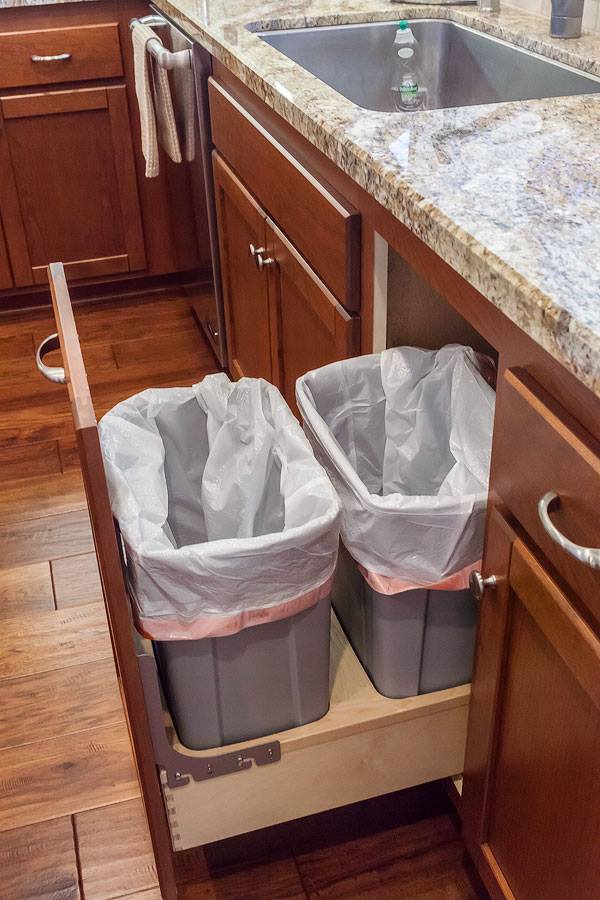 Pull-out kitchen trash and recycling bins beside a granite countertop and sink, with wooden cabinets.