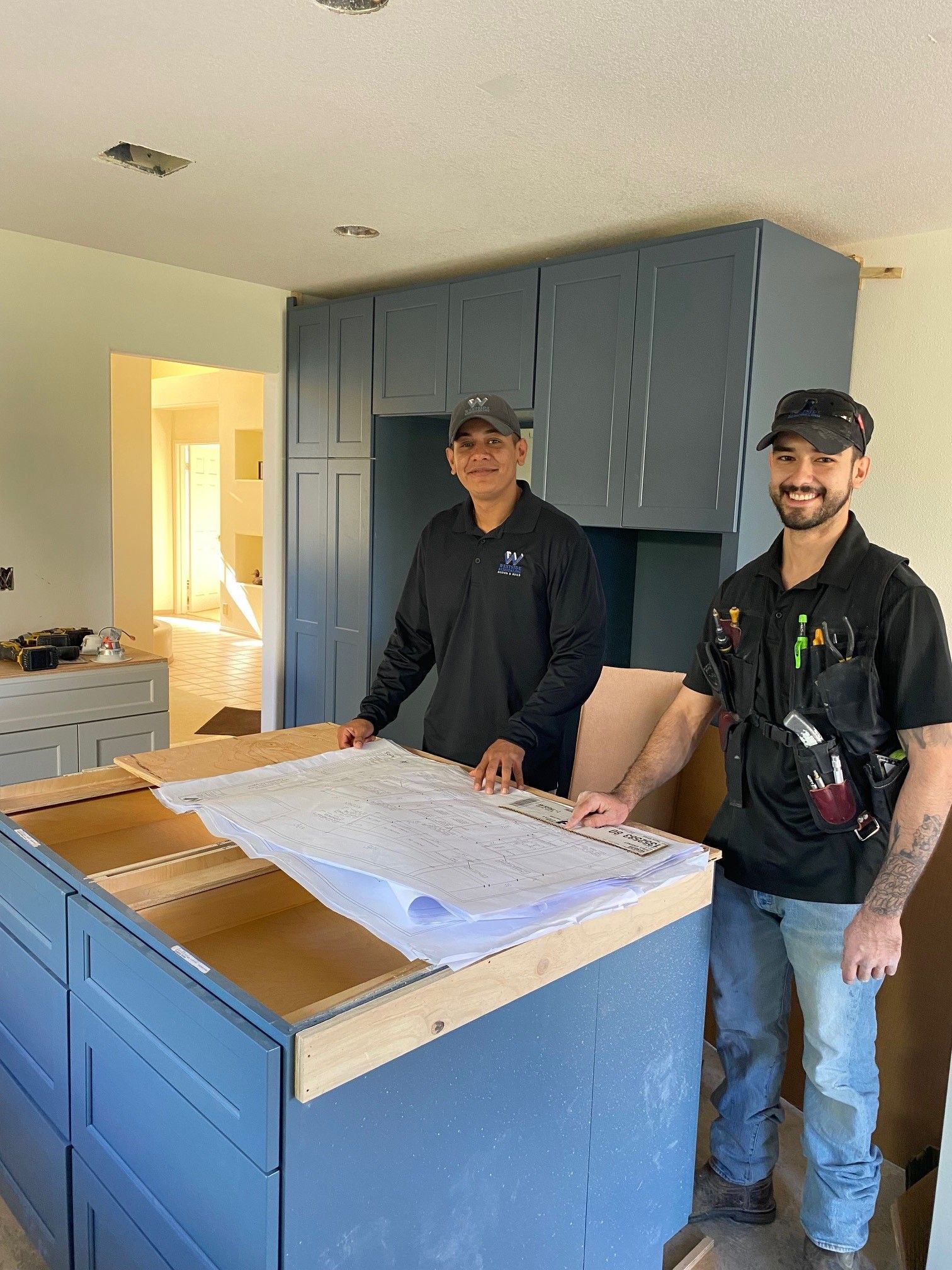 Two people standing behind a blue kitchen island with cabinets and a protective covering in a bright room