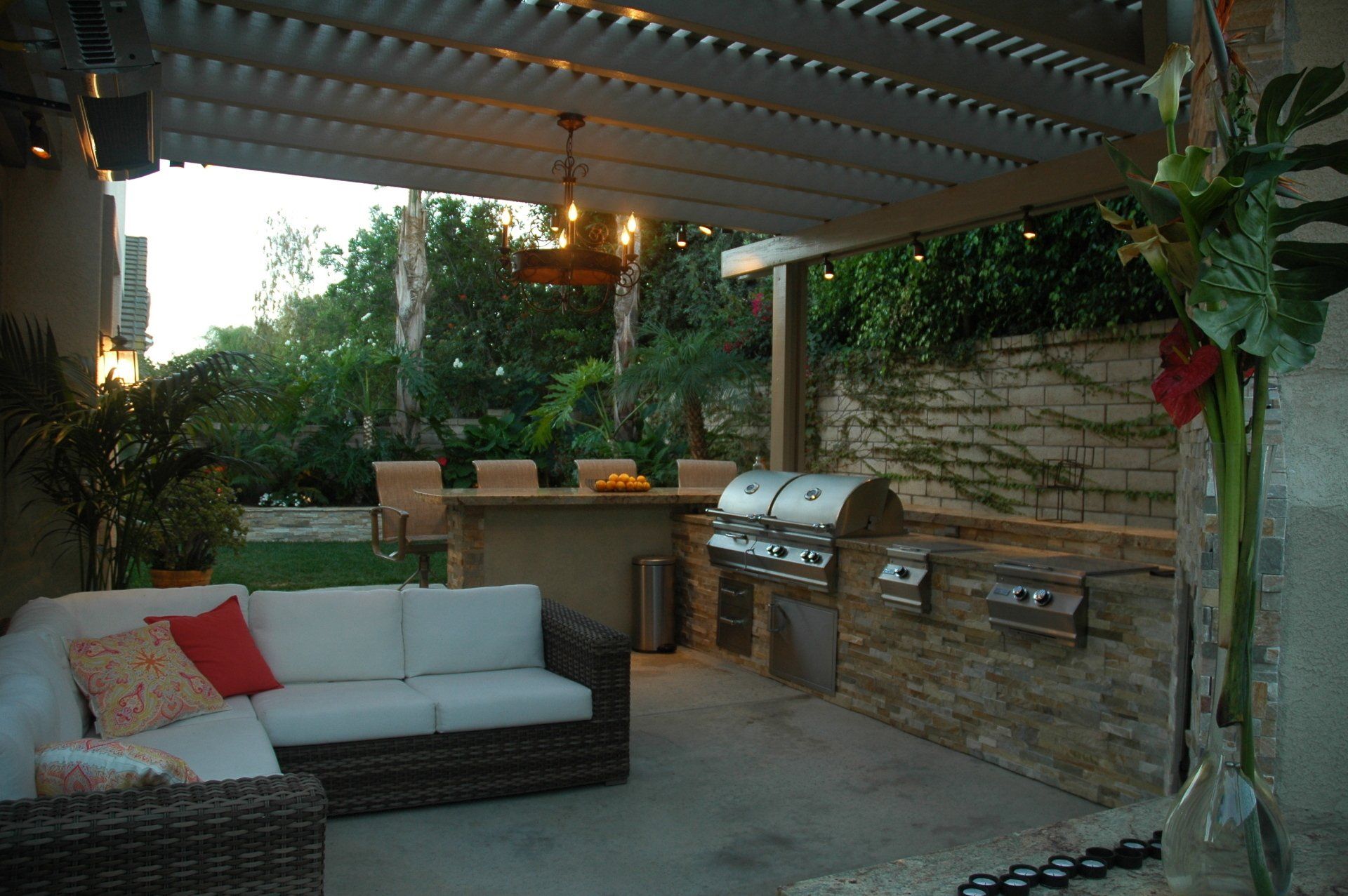 Covered patio with wicker sofa, grill, and hanging lights beside lush greenery at dusk