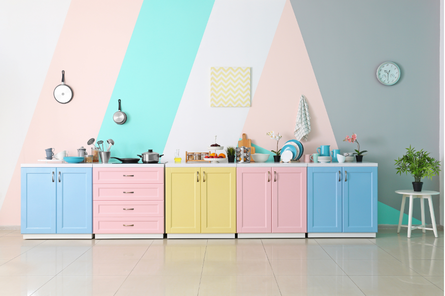 Pastel kitchen with colorful cabinets, utensils, wall clocks, and a potted plant on the right
