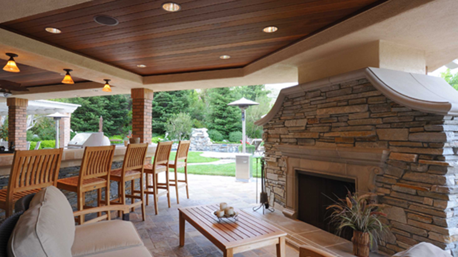 Covered outdoor patio with stone fireplace, wooden dining set, and cushioned seating overlooking a green lawn