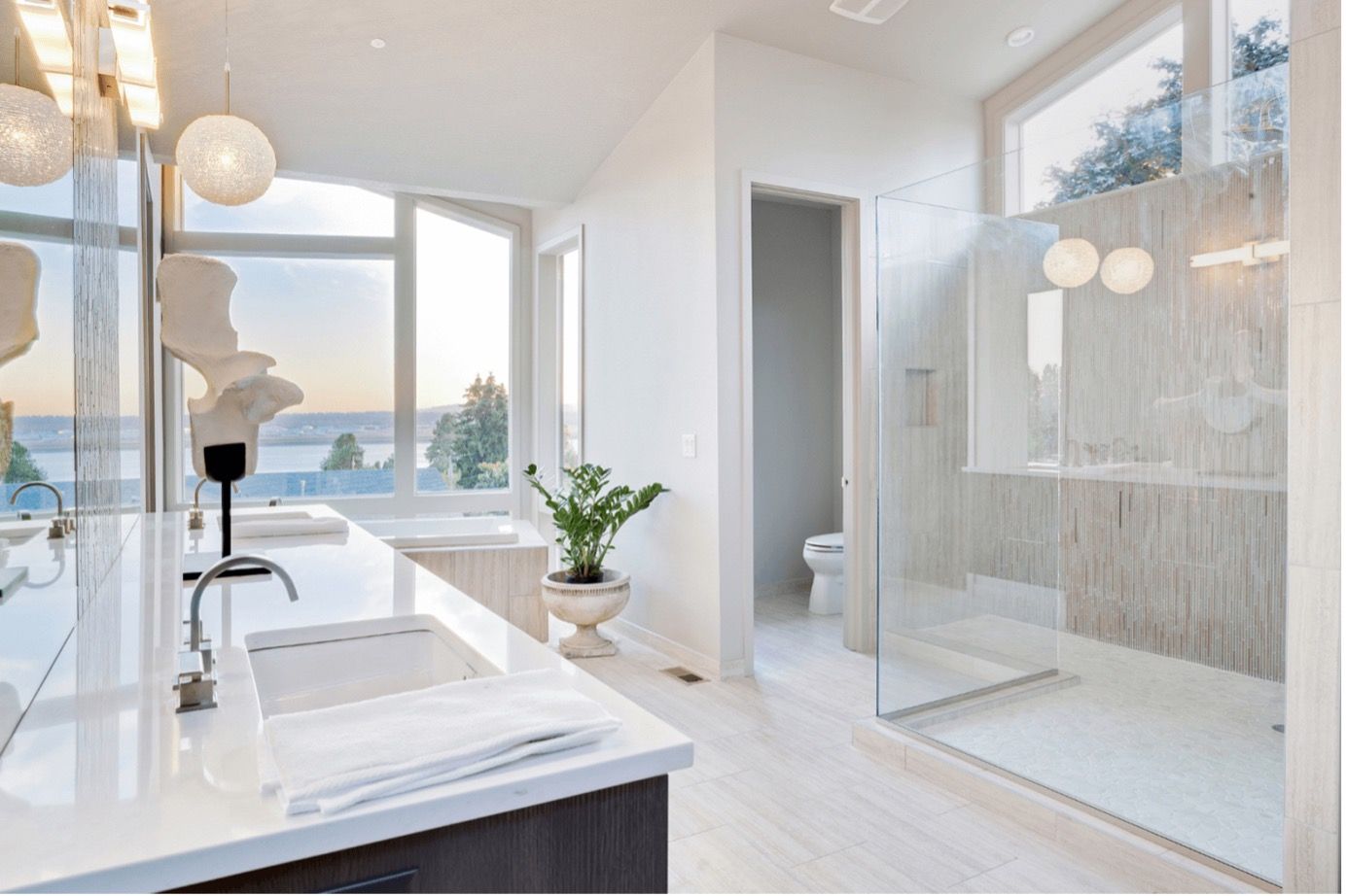 Bright modern bathroom with glass shower, marble vanity, potted plant, and ocean view windows