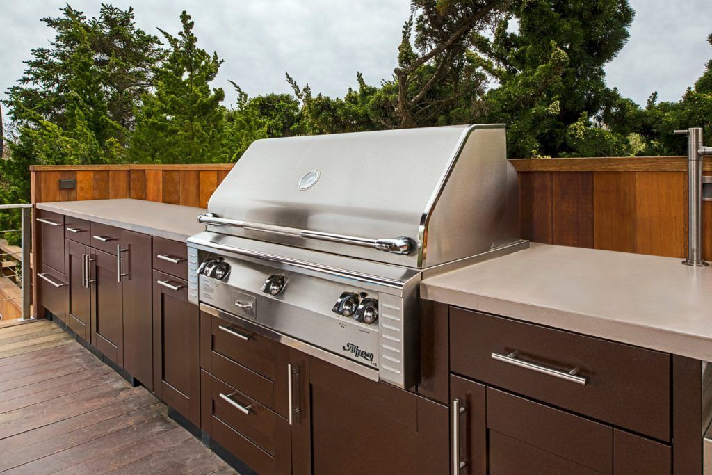 Outdoor stainless steel gas grill with brown cabinets on a wooden deck near a fence and trees