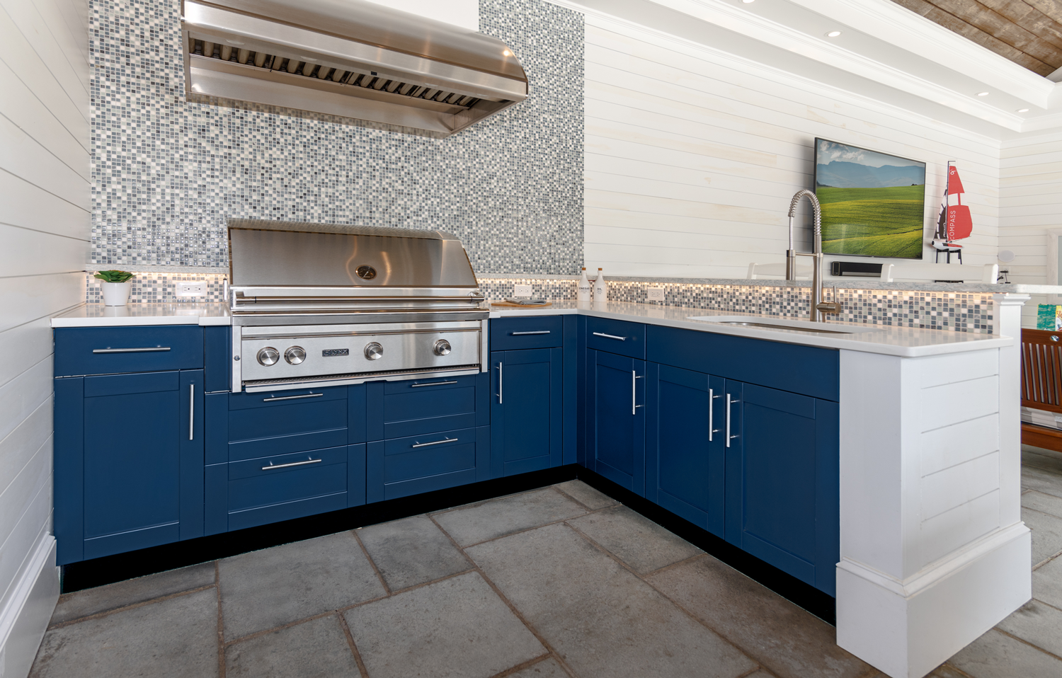 Blue outdoor kitchen with built-in grill, stainless hood, sink, and white countertop on stone patio
