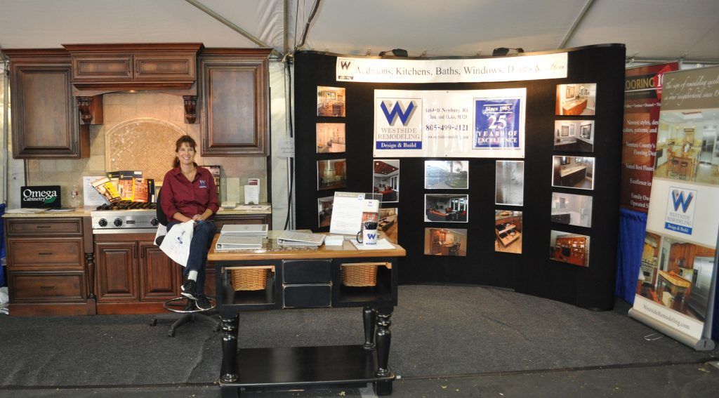 Trade show booth with wood cabinets, brochures, and a seated person at a display table under a tent.