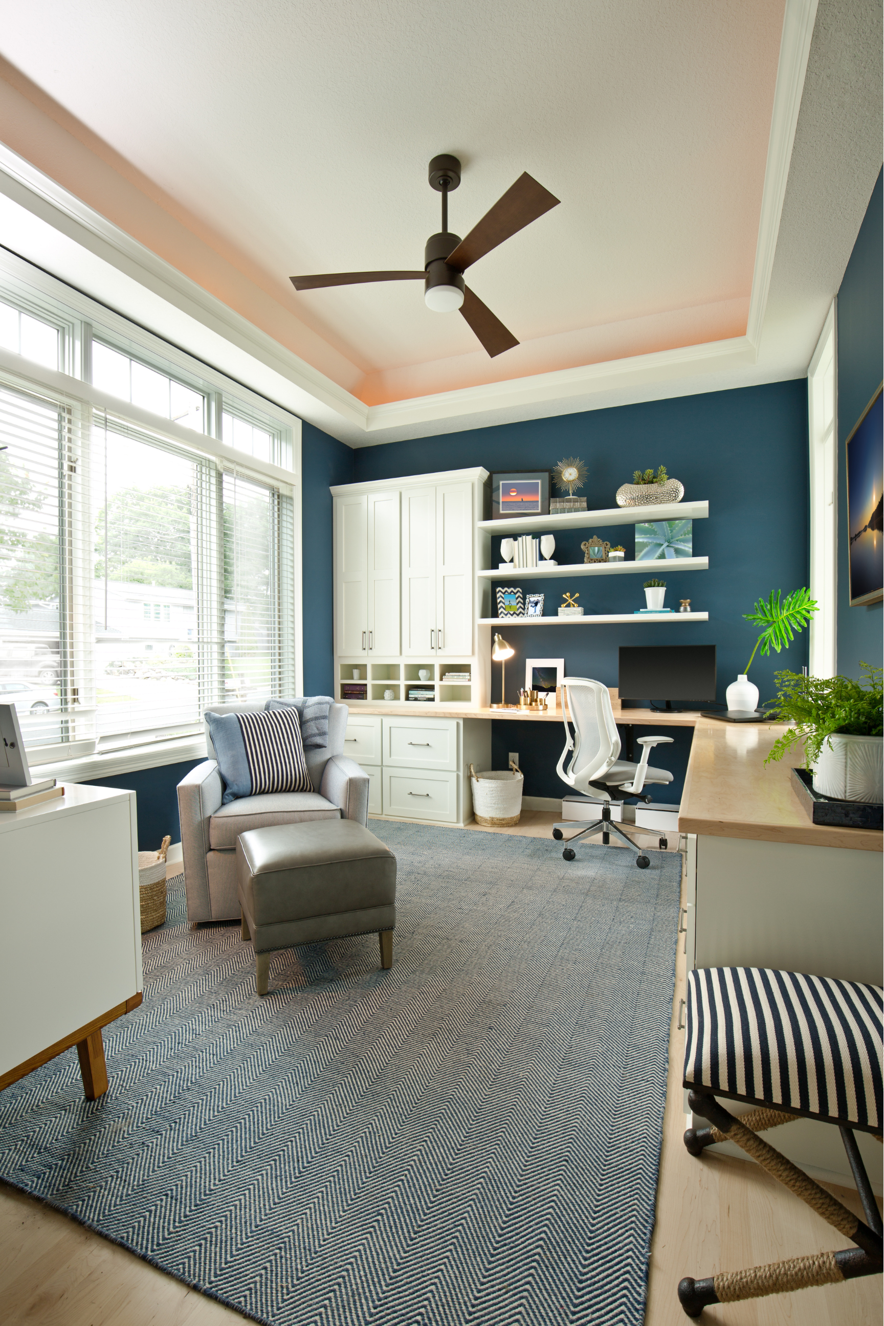 Bright home office with blue walls, white desk, chair, shelves, and large window with patterned rug.