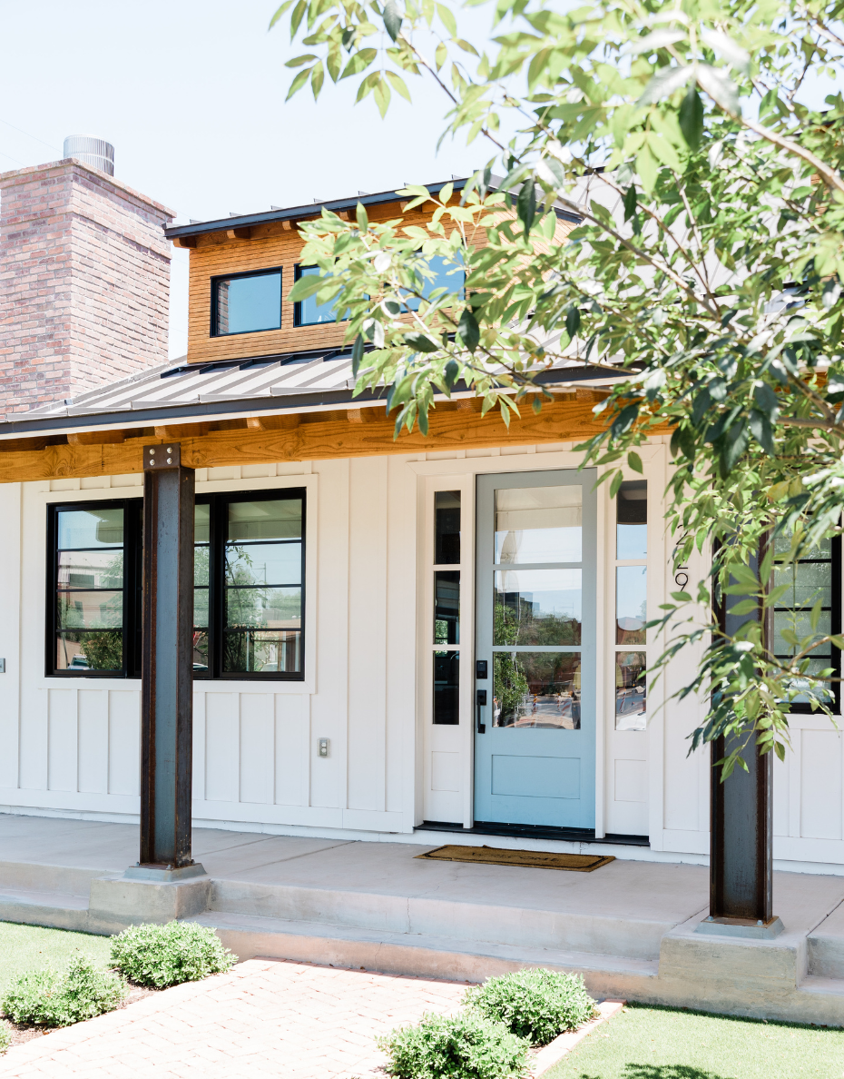 Modern white house facade with black trim, brick chimney, and glass doors under a leafy tree