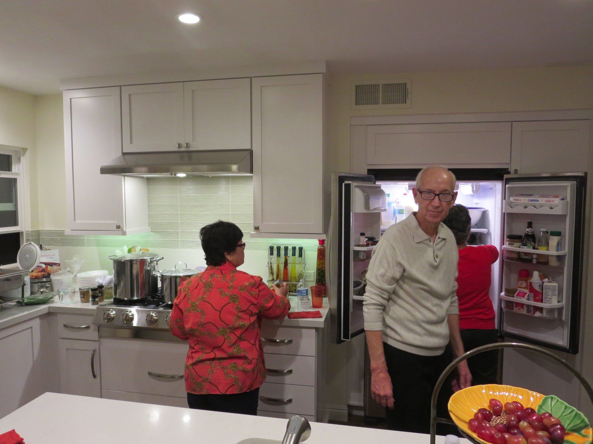 People in a bright kitchen, one at the stove and one by an open refrigerator.