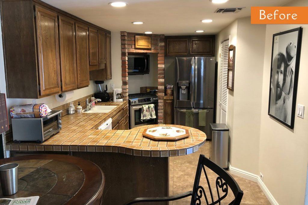 Before view of a warm kitchen with wood cabinets, granite counters, stainless fridge, and a patterned barstool.