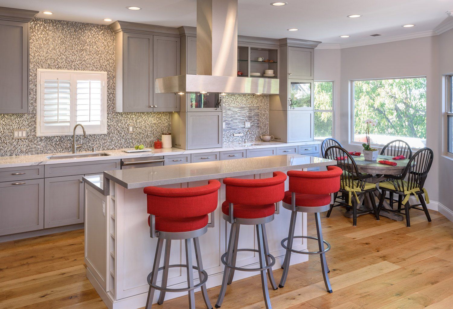 Bright modern kitchen with red barstools, stainless island, patterned backsplash, and dining nook by windows