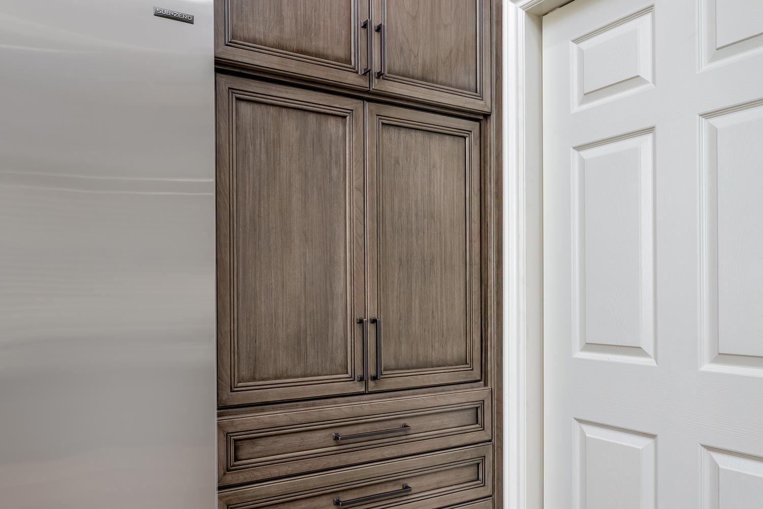 Built-in wooden pantry cabinet beside a white paneled door and stainless steel refrigerator