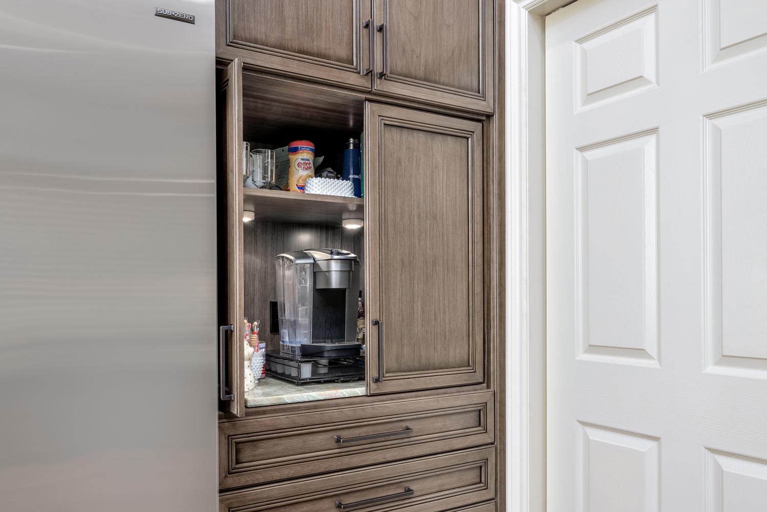 Kitchen cabinet with an open glass door showing a coffee maker and supplies inside