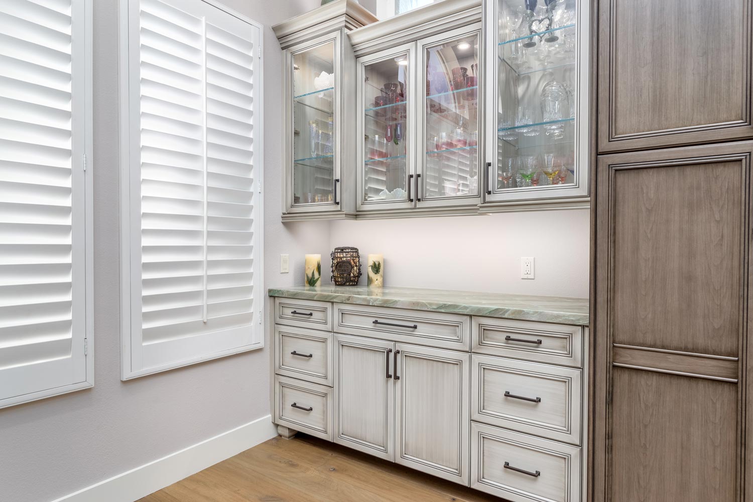 Bright built-in bathroom vanity with glass cabinets, white shutters, and a wood cabinet wall