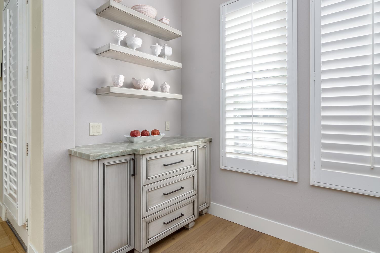 Bright white bathroom with window shutters, floating shelves, and a distressed vanity cabinet