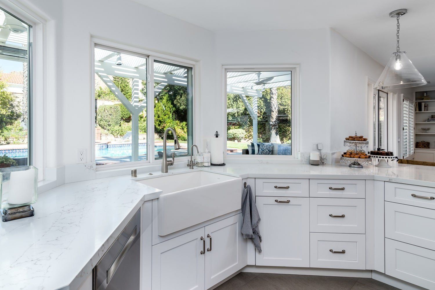 Bright white kitchen with large windows, farmhouse sink, marble counters, and a fruit stand on the island.