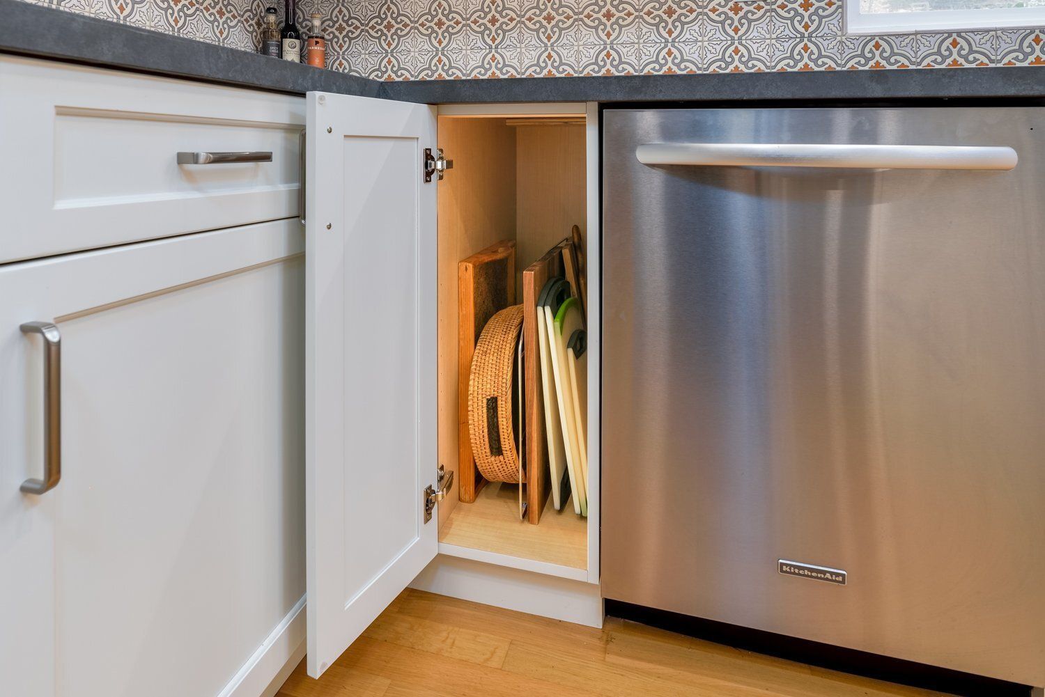 Open cabinet beside stainless dishwasher reveals pull-out tray organizers under a kitchen counter