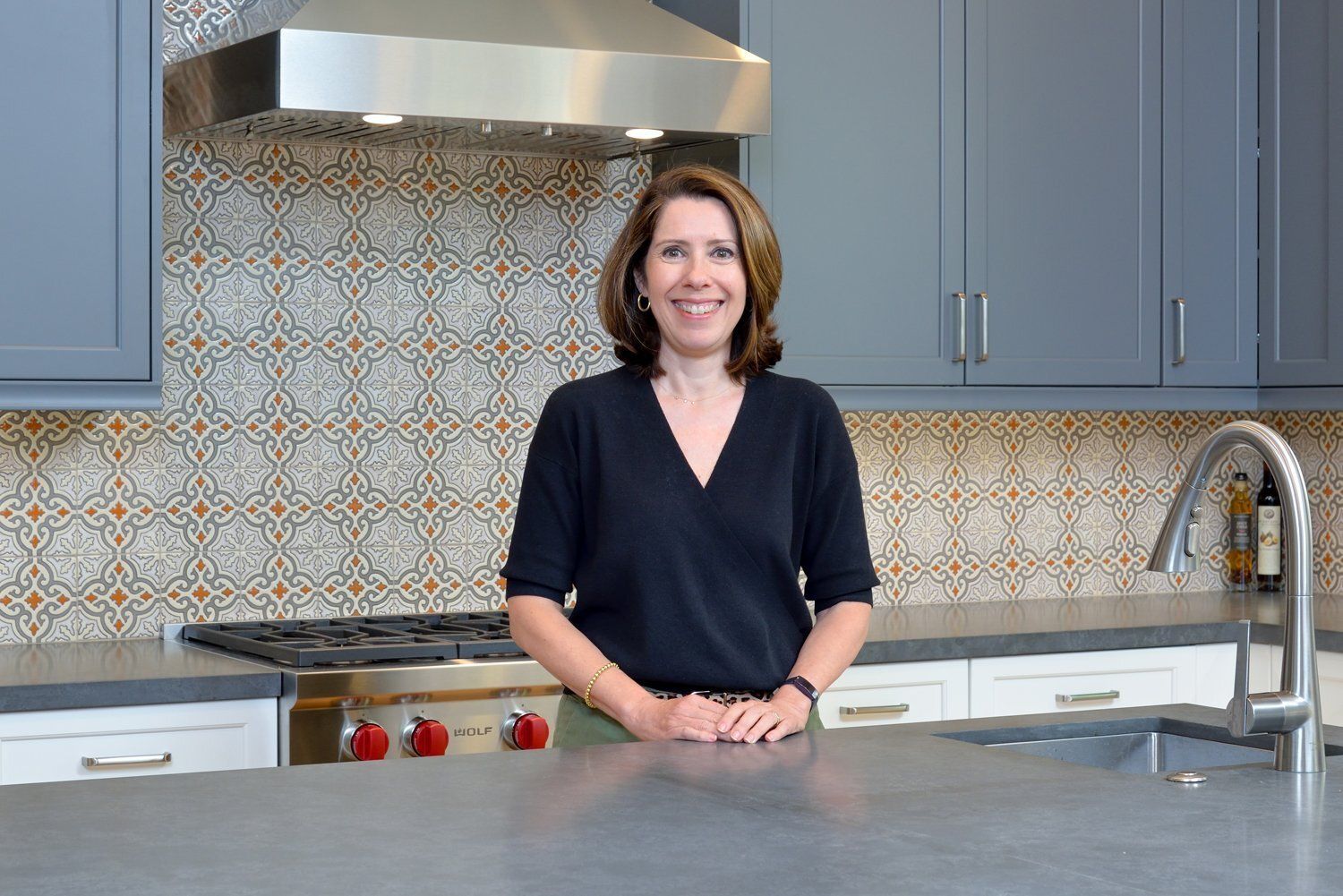 Woman standing in a modern kitchen behind a countertop and stove, smiling at the camera.