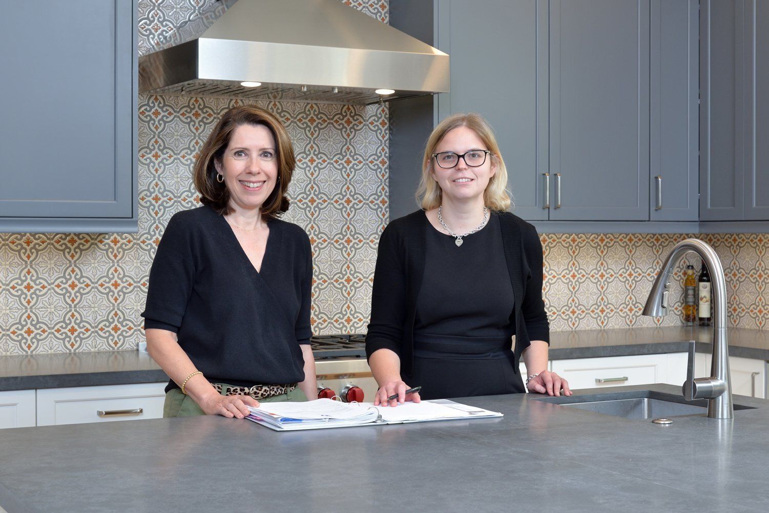 Two people standing in a modern kitchen behind a countertop, with a stainless steel range hood and patterned backsplash.