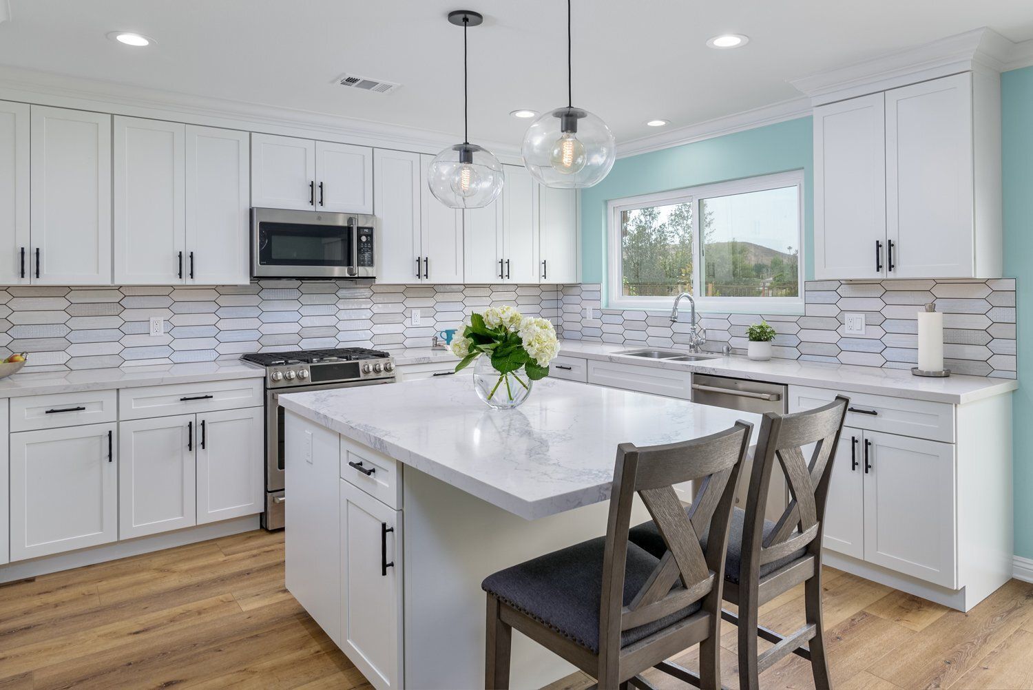 Bright white kitchen with marble island, wood floors, pendant lights, and two chairs by a window