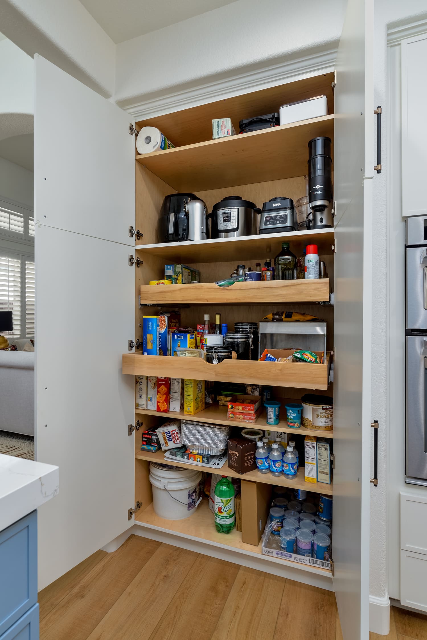 Open pantry cabinet with shelves of food, snacks, and small appliances in a kitchen.