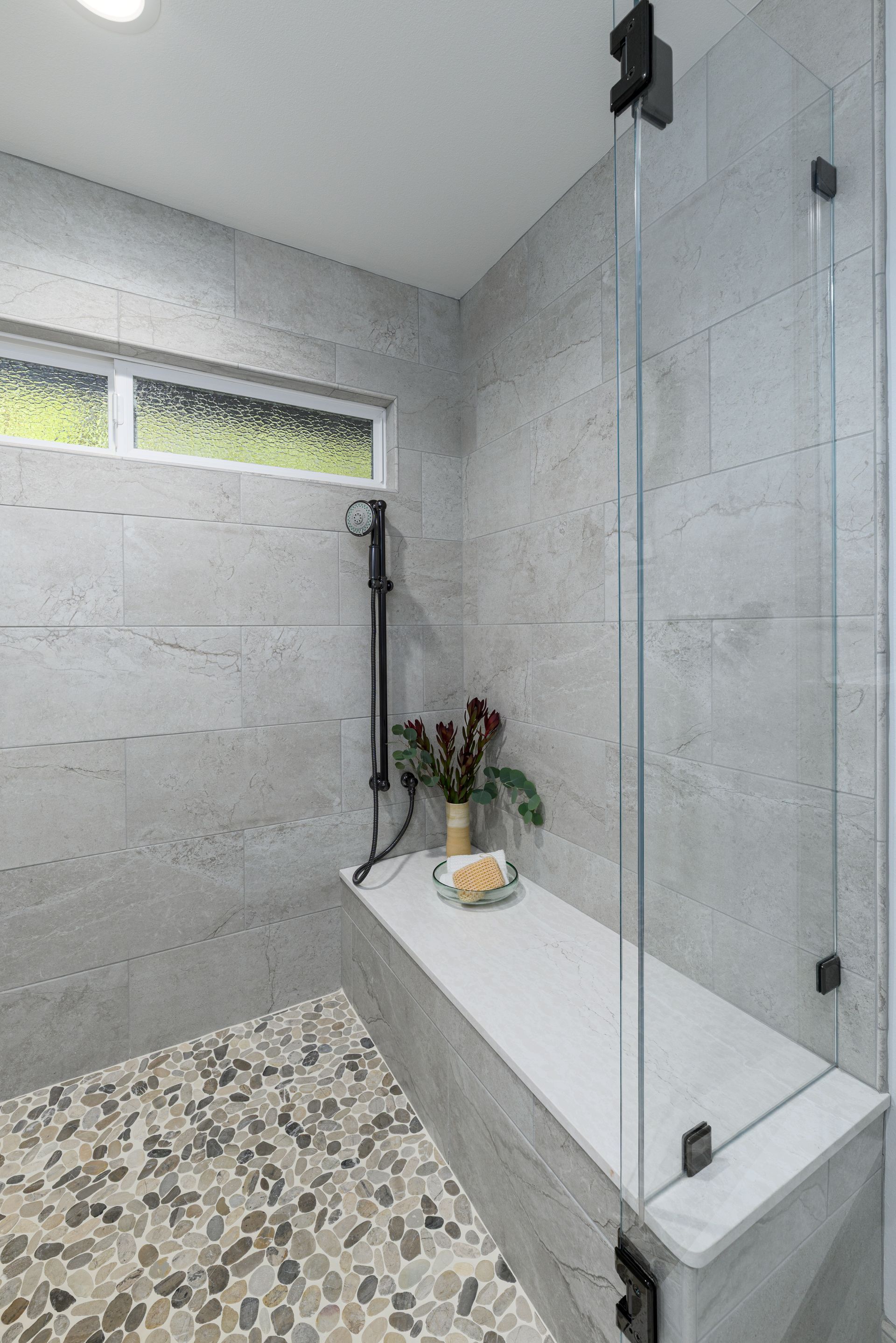 Modern gray tiled shower with a clear glass partition, white bench, and pebble floor.