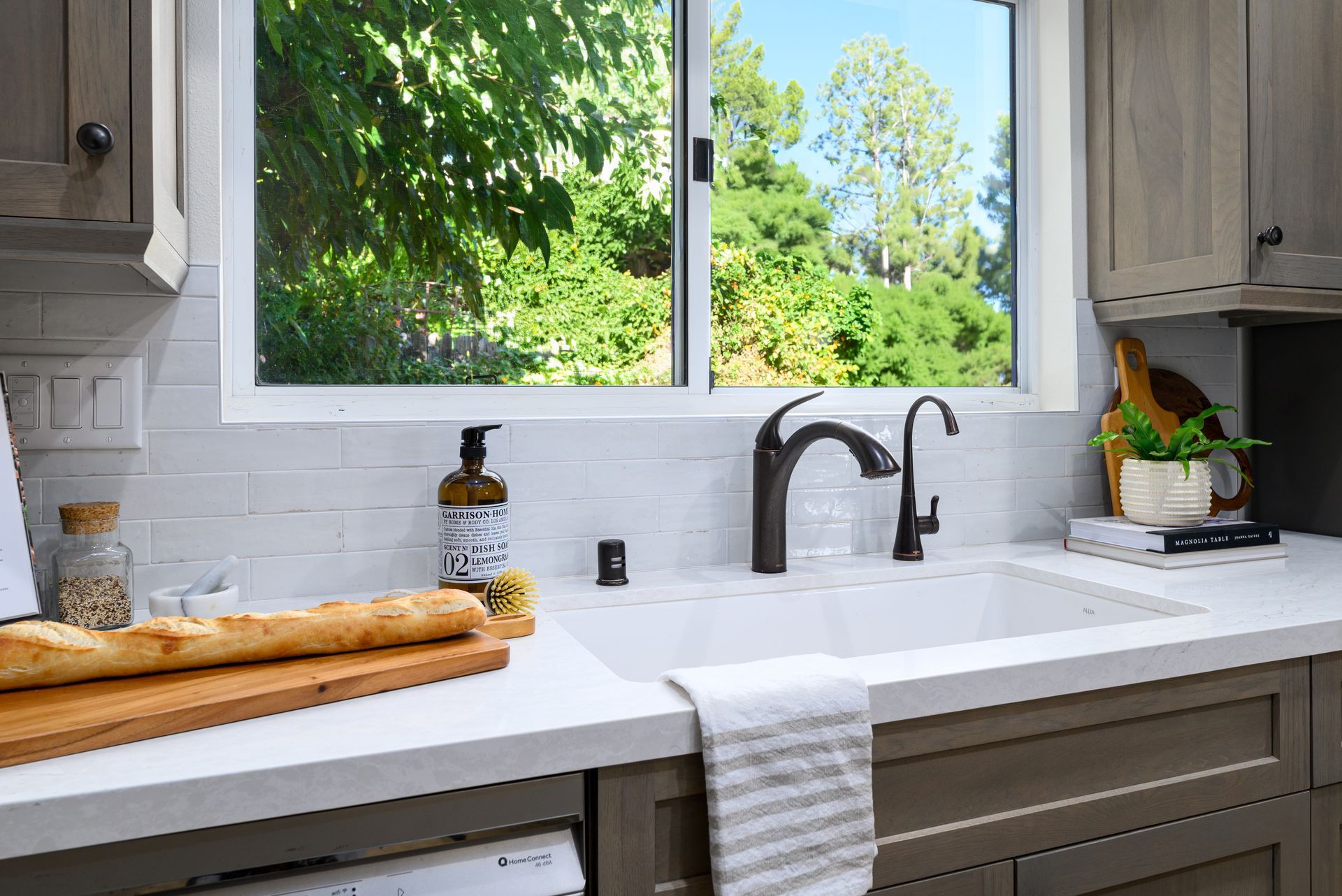 Bright kitchen sink under window with white countertop, soap dispenser, faucet, and wooden cutting board.