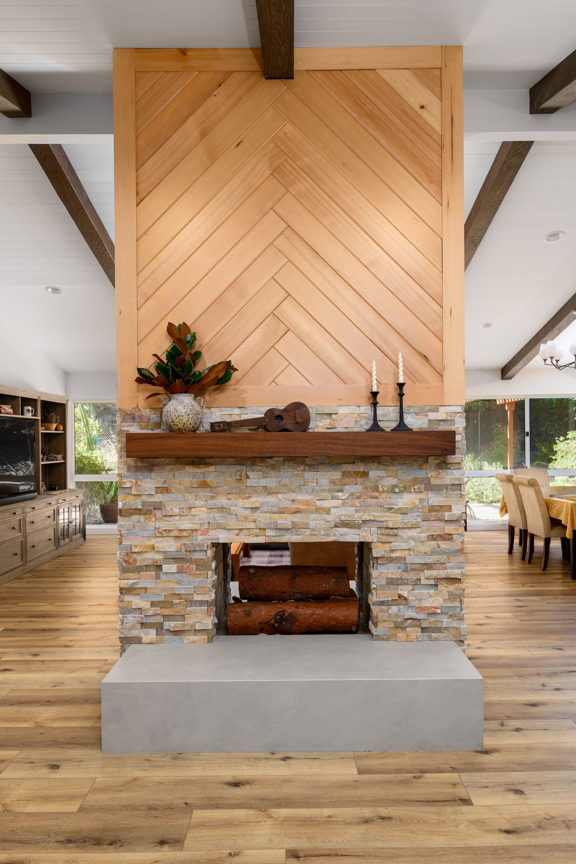 Modern rustic fireplace with stone surround, wood mantel, and herringbone wood paneling in a bright room