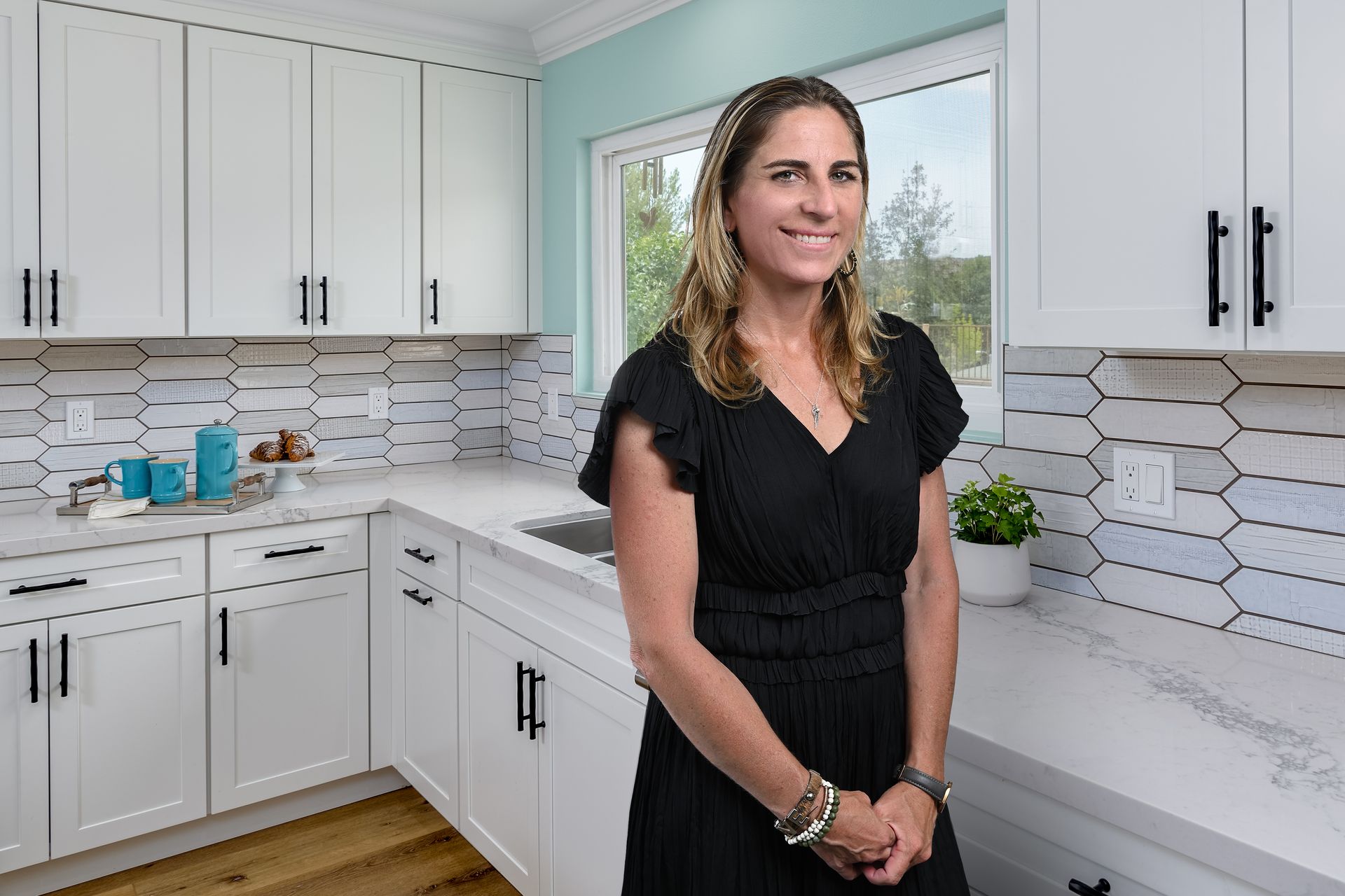 Woman standing in a bright white kitchen with hexagon tile backsplash and large window.