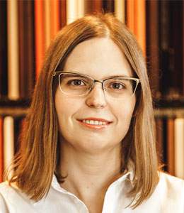 Portrait of a smiling person with glasses, straight light brown hair, and a white shirt, in front of bookshelves