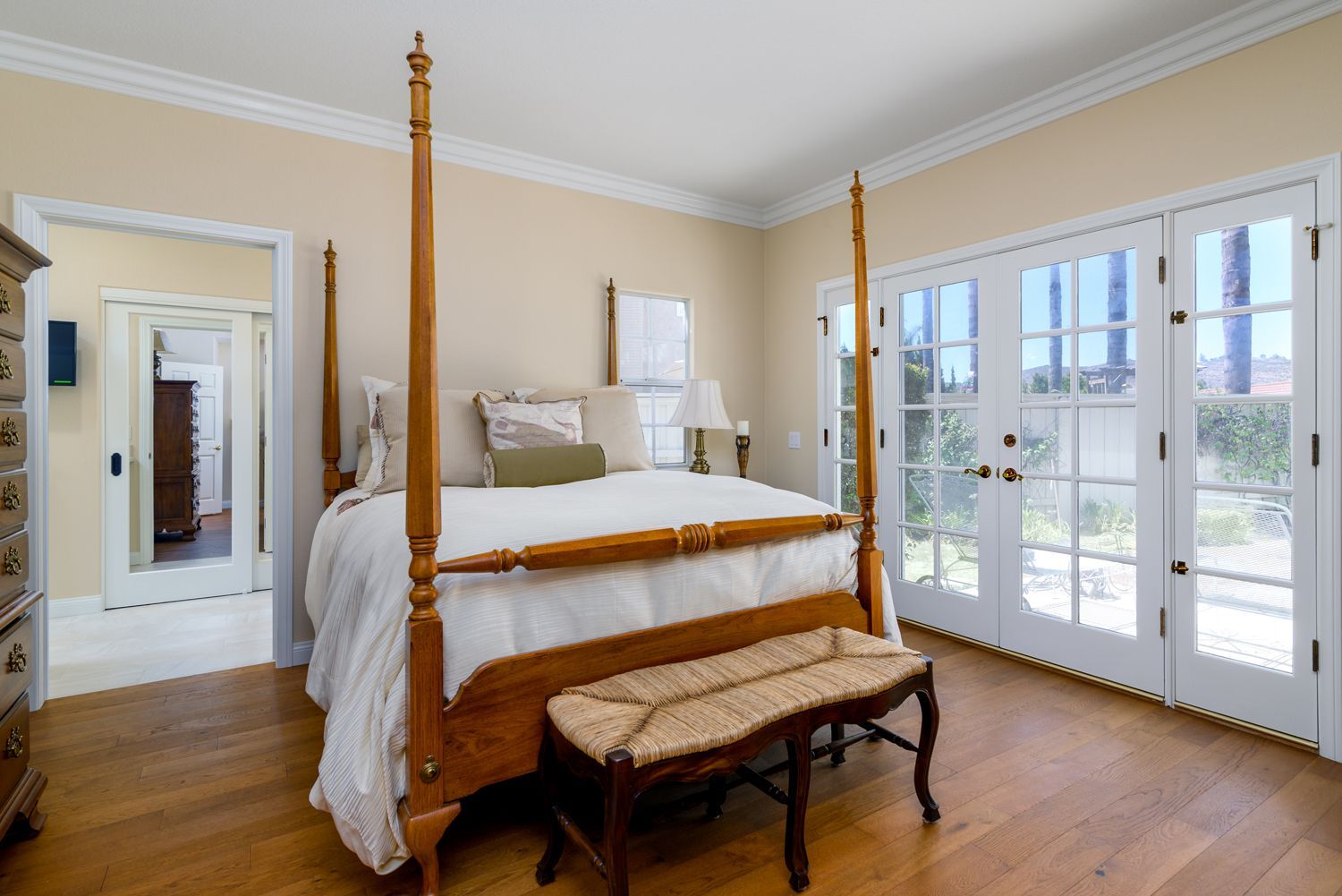 Bright bedroom with a four-poster bed, bench, and French doors opening to a balcony.