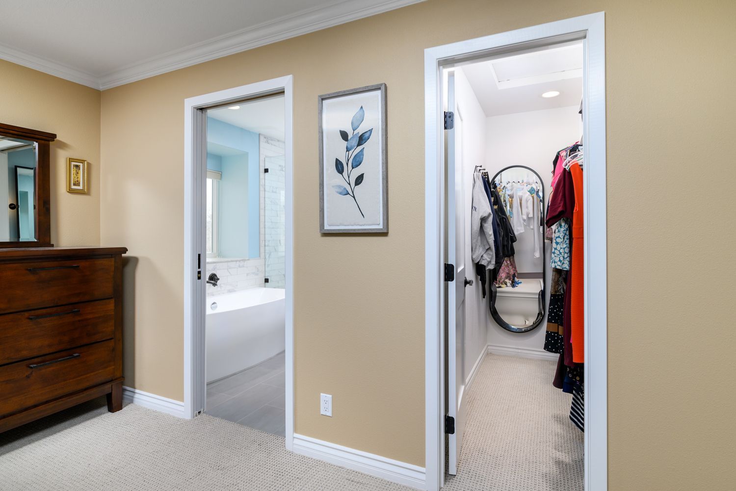 Bedroom hallway with open bathroom and closet doors, beige walls, dresser, and wall art