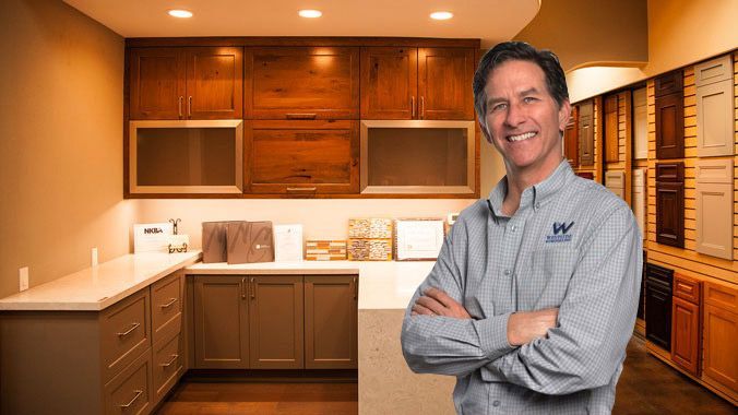 Smiling man in gray shirt standing in a warm-lit kitchen with wood cabinets and counters
