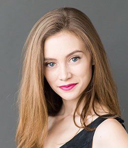 Portrait of a woman with long light brown hair, blue eyes, and pink lipstick, against a gray background