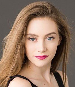 Portrait of a woman with long light brown hair, blue eyes, and bright pink lipstick against a gray background