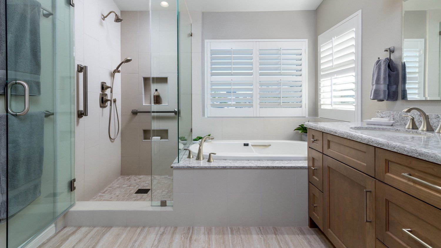 Accessible modern bathroom with glass shower, white tub, and wood vanity near a window with shutters